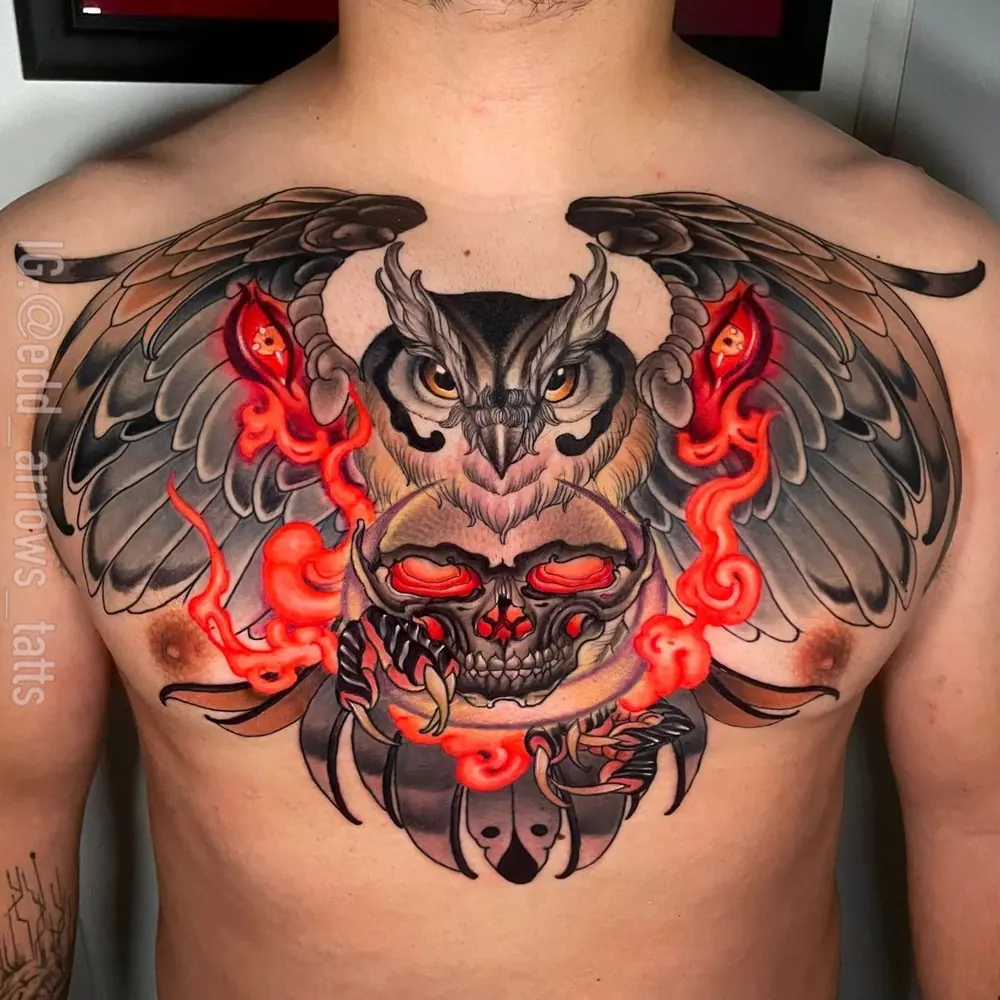 Chest tattoo: an owl with spread wings over a skull, with red flames, in neo-traditional color
